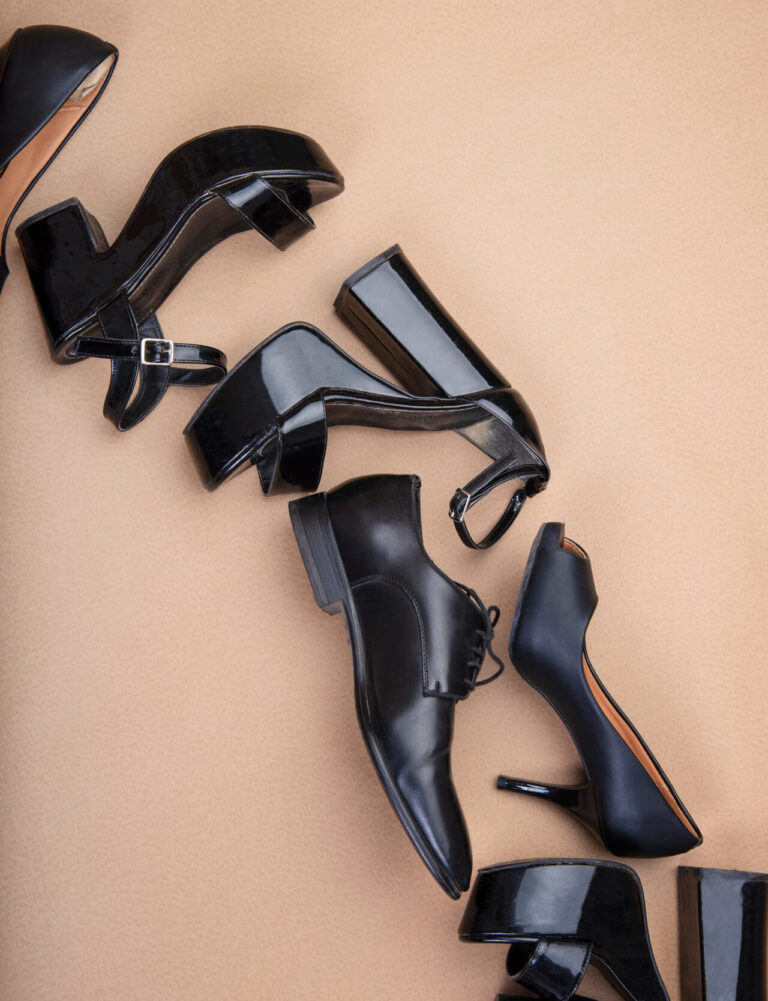 Composizione di scarpe eleganti nere — decolleté, sandali con tacco e scarpe derby — disposte su sfondo beige.