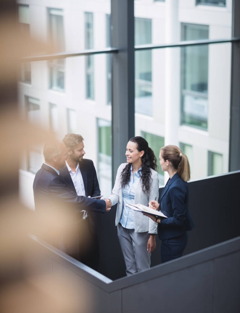 Quattro professionisti in abito formale si stringono la mano durante un incontro di lavoro in un ufficio con grandi vetrate.